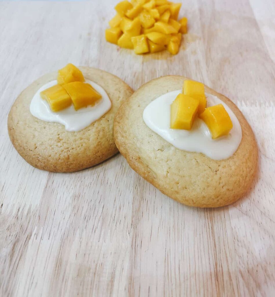 Bánh cookie mềm nhân xoài với lớp vỏ vàng và phần nhân xoài chảy, đặt trên nền pastel vàng, thương hiệu Mimi Bakes