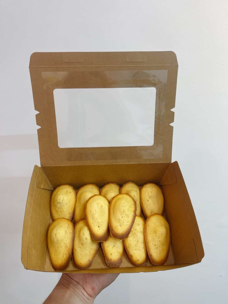 Hộp bánh với 12 madeleines vàng ruộm trong giấy lót, đặt trên nền màu pastel