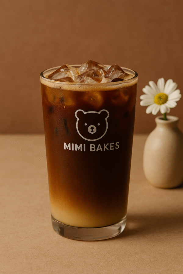 Ly cà phê sữa đá với hai tầng màu nâu đậm và sữa đặc, đá viên, logo Mimi Bakes, nền nâu pastel