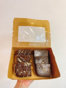 Hộp kraft vuông chứa 6 miếng brownie: 3 socola đen, 3 phủ dừa nạo, bên cạnh là hoa cúc trắng, nền màu nâu pastel